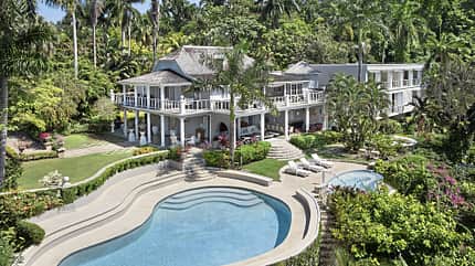 Villa Rio Chico, Luxusvilla, Ferienvilla, Swimmingpool, Tropengarten, Palmen, Außenansicht, Terrassenanlage, Architektur, Fly Unique Das Foto zeigt die Villa Rio Chico mit weitläufiger Gartenanlage und geschwungenem Swimmingpool in tropischer Umgebung. Die mehrteilige Anlage verfügt über umlaufende Veranden, weiße Balustraden, terrassierte Pool- und Liegeflächen sowie dichten Bewuchs mit Palmen. Die Aufnahme ist eine helle Außenansicht bei Tageslicht aus erhöhter Perspektive.