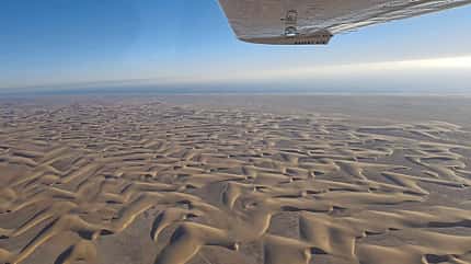 Desert Air, Fly Unique, Rundflug, Kleinflugzeug, Luftaufnahme, Wüstenlandschaft, Sanddüne, Flugzeugflügel, Luftfahrt Die Aufnahme zeigt eine Wüstenlandschaft mit langgezogenen, windgeformten Sanddünen; im oberen Bildbereich ist die Unterseite eines Flugzeugflügels mit der Aufschrift „DESERT AIR“ sichtbar. Laut Dateiname handelt es sich um einen Flug von Desert Air (Fly Unique), aufgenommen aus der Luft in schräger Perspektive. Das Bild datiert dem Dateinamen zufolge auf den 24.09.2026.