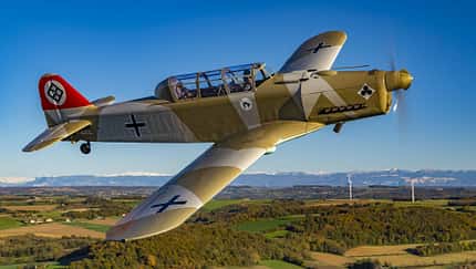 Pilatus P2, Pilatus-Trainingsflugzeug, Propellertrainer, Oldtimerflugzeug, Warbird, Tandemsitzer, Eindecker, Luftaufnahme, Allgemeine Luftfahrt Das Foto zeigt eine Pilatus P2 im Flug. Der zweisitzige, einmotorige Propellertrainer mit Tandemcockpit ist in Tarnlackierung mit historischen Hoheitszeichen zu sehen; der laufende Propeller und die verglaste Haube sind deutlich erkennbar. Die Aufnahme ist eine Luftaufnahme über ländlicher Landschaft mit Gebirgskette im Hintergrund.