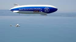 Technoflug Piccolo und Zeppelin NT in der Luft über einem See, vermutlich Bodensee, aus der Luft fotografiert. Das Luftschiff trägt die Aufschrift 'Next Generation Mobility. ZF NOW.' und ein einmotoriges Leichtflugzeug (Technoflug Piccolo) fliegt darunter.