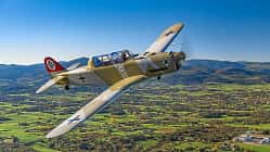 Das Foto zeigt eine Pilatus P2 im Flug in schräger Seitenansicht über einer ländlichen Landschaft mit Hügelkette im Hintergrund. Der einmotorige Tiefdecker mit Tandemsitz-Kabine und Propeller in Bewegung trägt eine historische Militärlackierung; typische Merkmale sind Kolbenmotor, Einziehfahrwerk und Spornrad. Die Aufnahme wirkt als Luft‑Luft‑Fotografie mit sichtbarem Cockpit und Pilot.