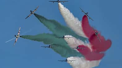 Mehrere Kunstflugzeuge fliegen in Formation und erzeugen farbige Rauchspuren in Grün, Weiß und Rot am Himmel.