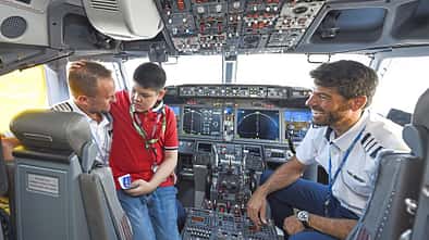 Das Bild zeigt das Cockpit eines Flugzeugs mit zwei Piloten und einem Kind, das auf dem Schoß eines Erwachsenen sitzt. Alle Personen wirken fröhlich und unterhalten sich.