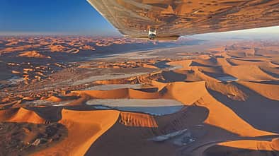 Luftaufnahme der Sossusvlei‑Wüste in Namibia, fotografiert aus einem Kleinflugzeug; der Flügel ist im oberen Bildbereich sichtbar. Die Szene zeigt weitläufige orangefarbene Sanddünen mit einzelnen ausgetrockneten Pfannen (u. a. Dead Vlei) und starkem Schattenwurf. Die Aufnahme entstand im Rahmen eines Rundflugs (Fly Unique), laut IPTC mit Hinweis auf eine Cessna als Trägerflugzeug.
