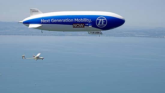 Technoflug Piccolo und Zeppelin NT in der Luft über einem See, vermutlich Bodensee, aus der Luft fotografiert. Das Luftschiff trägt die Aufschrift 'Next Generation Mobility. ZF NOW.' und ein einmotoriges Leichtflugzeug (Technoflug Piccolo) fliegt darunter.