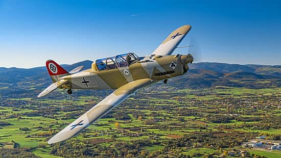 Das Foto zeigt eine Pilatus P2 im Flug in schräger Seitenansicht über einer ländlichen Landschaft mit Hügelkette im Hintergrund. Der einmotorige Tiefdecker mit Tandemsitz-Kabine und Propeller in Bewegung trägt eine historische Militärlackierung; typische Merkmale sind Kolbenmotor, Einziehfahrwerk und Spornrad. Die Aufnahme wirkt als Luft‑Luft‑Fotografie mit sichtbarem Cockpit und Pilot.