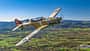 Das Foto zeigt eine Pilatus P2 im Flug in schräger Seitenansicht über einer ländlichen Landschaft mit Hügelkette im Hintergrund. Der einmotorige Tiefdecker mit Tandemsitz-Kabine und Propeller in Bewegung trägt eine historische Militärlackierung; typische Merkmale sind Kolbenmotor, Einziehfahrwerk und Spornrad. Die Aufnahme wirkt als Luft‑Luft‑Fotografie mit sichtbarem Cockpit und Pilot.