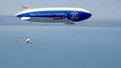 Technoflug Piccolo und Zeppelin NT in der Luft über einem See, vermutlich Bodensee, aus der Luft fotografiert. Das Luftschiff trägt die Aufschrift 'Next Generation Mobility. ZF NOW.' und ein einmotoriges Leichtflugzeug (Technoflug Piccolo) fliegt darunter.
