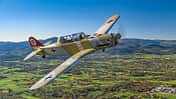 Das Foto zeigt eine Pilatus P2 im Flug in schräger Seitenansicht über einer ländlichen Landschaft mit Hügelkette im Hintergrund. Der einmotorige Tiefdecker mit Tandemsitz-Kabine und Propeller in Bewegung trägt eine historische Militärlackierung; typische Merkmale sind Kolbenmotor, Einziehfahrwerk und Spornrad. Die Aufnahme wirkt als Luft‑Luft‑Fotografie mit sichtbarem Cockpit und Pilot.