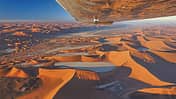 Luftaufnahme der Sossusvlei‑Wüste in Namibia, fotografiert aus einem Kleinflugzeug; der Flügel ist im oberen Bildbereich sichtbar. Die Szene zeigt weitläufige orangefarbene Sanddünen mit einzelnen ausgetrockneten Pfannen (u. a. Dead Vlei) und starkem Schattenwurf. Die Aufnahme entstand im Rahmen eines Rundflugs (Fly Unique), laut IPTC mit Hinweis auf eine Cessna als Trägerflugzeug.
