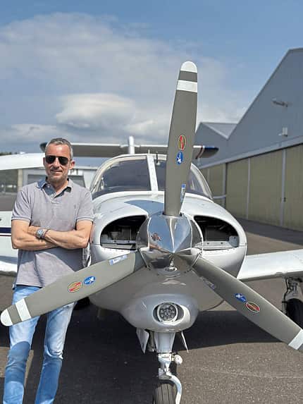 Piper PA-28, Piper-Leichtflugzeug, Einmotoriges Propellerflugzeug, Schulungsflugzeug, Allgemeine Luftfahrt, Dreiblattpropeller, Nasenradfahrwerk, Kolbentriebwerk, Flugplatz, Cockpitkanzel Das Foto zeigt eine Piper PA-28, ein einmotoriges Leichtflugzeug, in Frontalansicht am Boden. Der Dreiblattpropeller mit polierter Spinnerkappe, das Nasenradfahrwerk und die Tiefdecker-Bauweise sind deutlich erkennbar. Neben der Flugzeugnase steht eine Person; im Hintergrund befinden sich Hangars eines Flugplatzes.